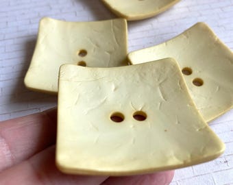 May include: Four square, pale yellow buttons with a slightly concave surface and a textured, crackled finish. Each button features two centered holes for attachment, arranged horizontally in the middle of the square shape.