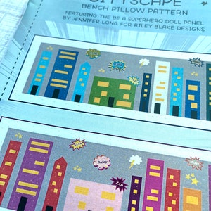 May include: A pattern for a cityscape bench pillow featuring a superhero doll panel. The pattern includes a variety of buildings with colorful accents and text like "Super", "Bang!" and "Zap!"