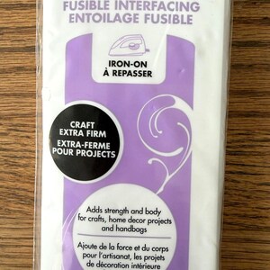 Op de afbeelding: Een verpakking HeatnBond strijkbare tussenvoering in een witte en paarse verpakking. Het label vermeldt HeatnBond, Fusible Interfacing, Iron-On en Craft Extra Firm. Het voegt stevigheid en vorm toe voor knutselwerk en handtassen.