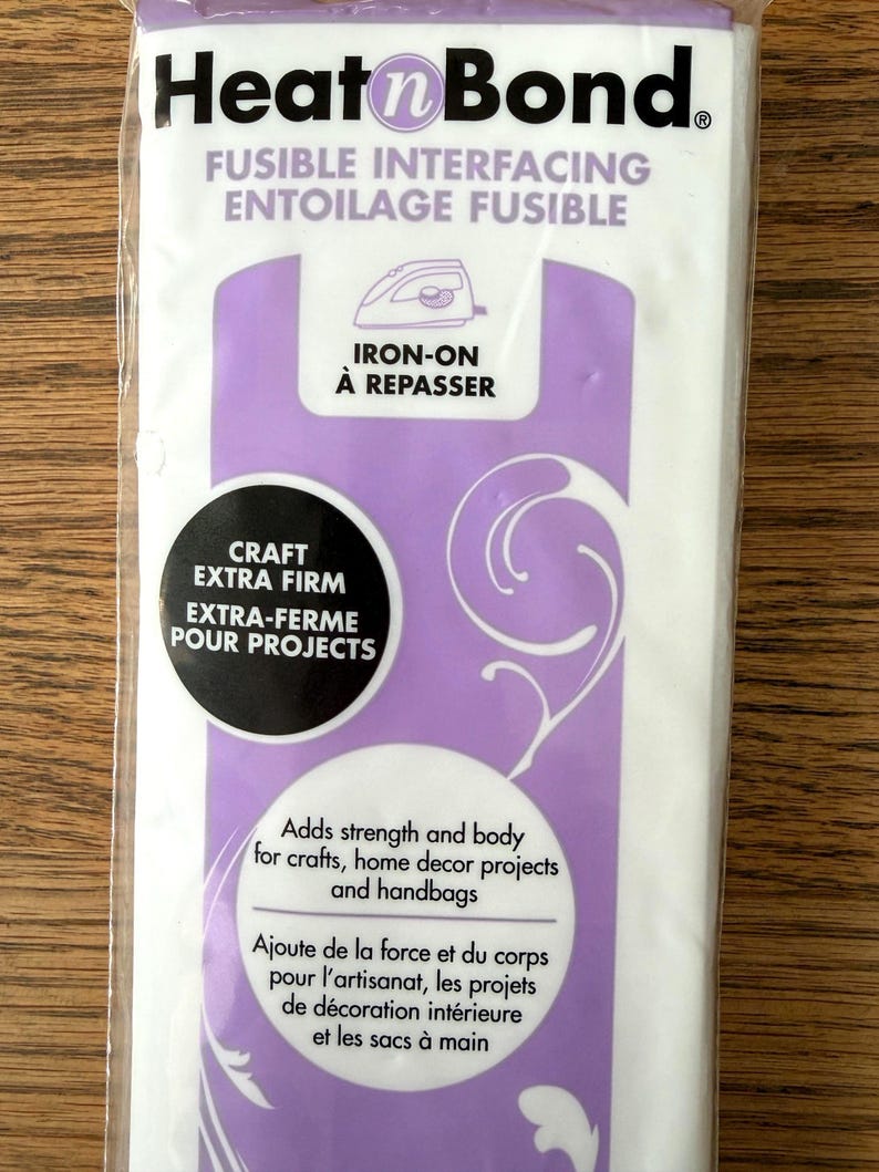 Op de afbeelding: Een verpakking HeatnBond strijkbare tussenvoering in een witte en paarse verpakking. Het label vermeldt HeatnBond, Fusible Interfacing, Iron-On en Craft Extra Firm. Het voegt stevigheid en vorm toe voor knutselwerk en handtassen.