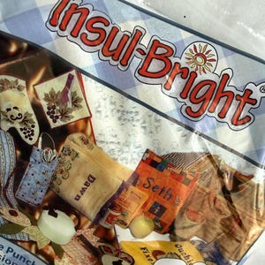 May include: A package of Insul-Bright insulated lining featuring a blue and white checkered background. The packaging displays various quilted items including a mitt, bags with names like Dawn and Seth, and a free sewing pattern label in English.