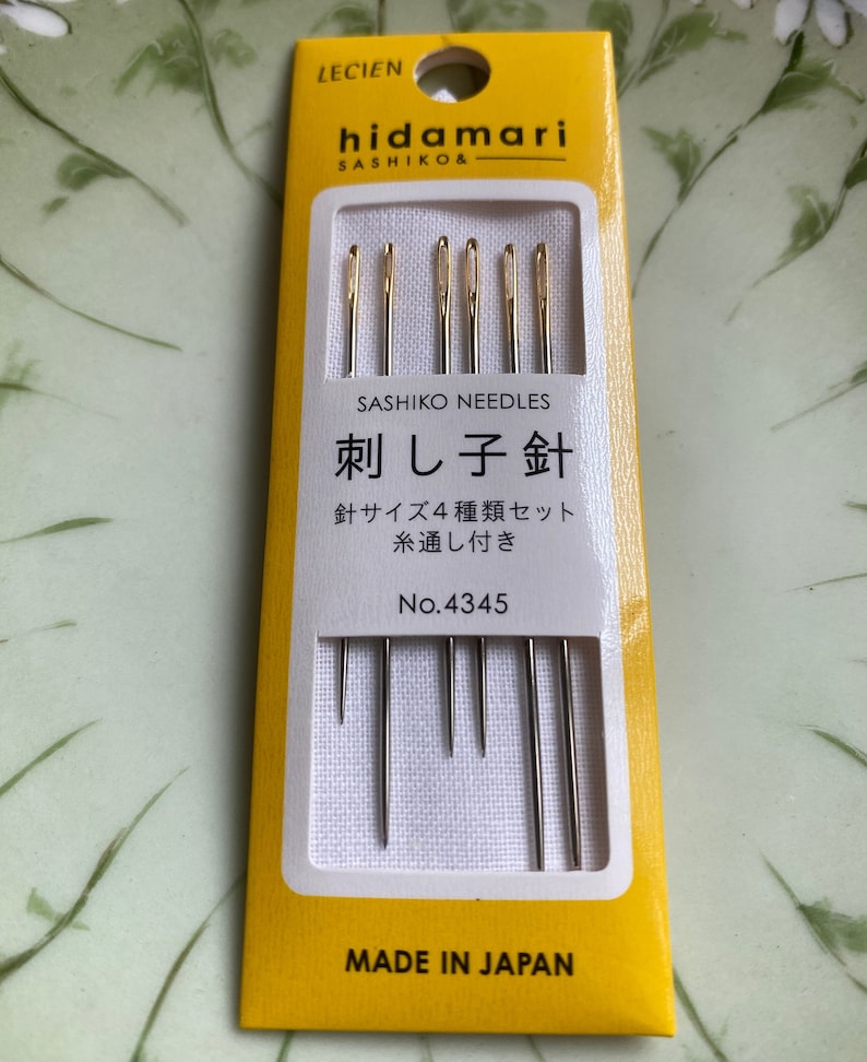 May include: A yellow and white package of sashiko needles. The package has the brand name "hidamari" and the text "SASHIKO NEEDLES" printed on it. The package contains several needles with gold-colored eyes. The text "MADE IN JAPAN" is printed at the bottom.