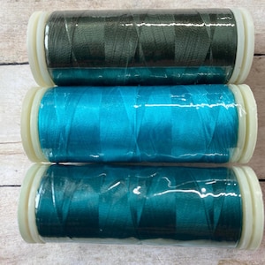 May include: Three spools of thread in shades of teal, turquoise, and dark green. The thread is wrapped in clear plastic and has white plastic ends. The spools are stacked vertically, showcasing the vibrant colors.