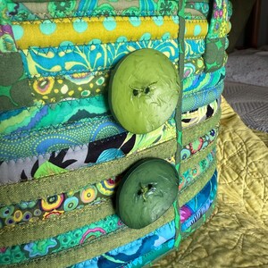 May include: A fabric basket constructed from coiled strips of multi-colored patterned cotton fabric in shades of green, blue, and yellow. The basket features two large, round, olive green buttons attached to the front and two fabric handles.