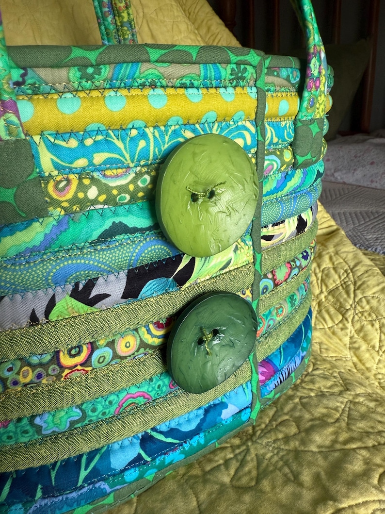May include: A fabric basket constructed from coiled strips of multi-colored patterned cotton fabric in shades of green, blue, and yellow. The basket features two large, round, olive green buttons attached to the front and two fabric handles.