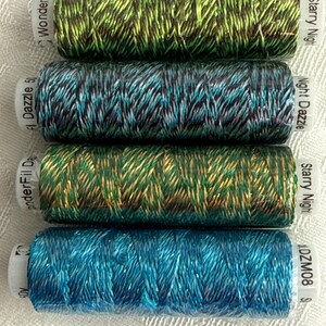 May include: Four spools of WonderFil Dazzle thread stacked horizontally. The top spool is green and black, the second is teal and black, the third is gold and green, and the bottom is bright blue. Each spool features metallic fibers.