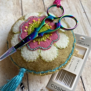 Pode incluir: Um par de tesouras de bordar multicoloridas repousa sobre uma almofada redonda com um design floral. A almofada apresenta um design floral rosa e branco com detalhes dourados e turquesas. Um pacote de agulhas tamb&eacute;m est&aacute; presente.