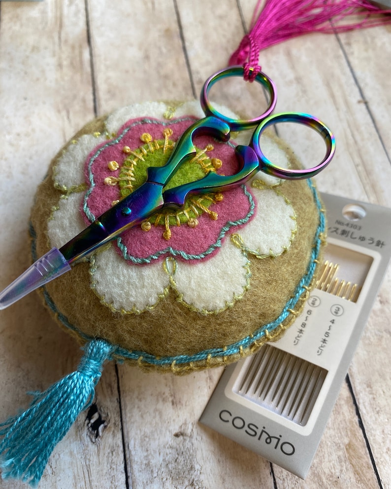 Pode incluir: Um par de tesouras de bordar multicoloridas repousa sobre uma almofada redonda com um design floral. A almofada apresenta um design floral rosa e branco com detalhes dourados e turquesas. Um pacote de agulhas tamb&eacute;m est&aacute; presente.
