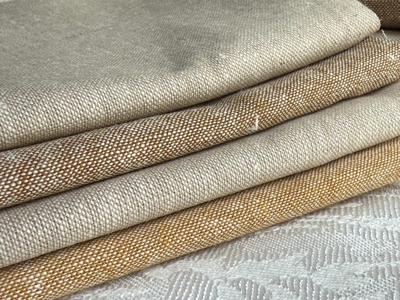 K&ouml;nnte beinhalten: Ein Stapel aus vier gefalteten Stoffst&uuml;cken mit einer sichtbaren Webstruktur. Die oberste Schicht ist hellbeige, gefolgt von einem mittleren Braunton, einem dunkleren Goldbraun und einer untersten Schicht in einem satten Senfgelb.