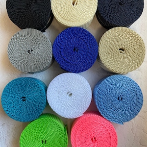 May include: A collection of twelve spools of woven ribbon in various colors, including black, cream, navy, gray, blue, tan, teal, white, light blue, green, and pink. The spools are tightly wound and arranged in a grid pattern, showcasing the range of available colors.