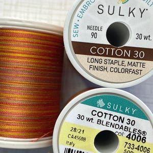 May include: Two spools of Sulky Cotton 30 thread. One spool shows variegated orange, yellow, and brown thread. The labels display text including SULKY, COTTON 30, LONG STAPLE, MATTE FINISH, COLORFAST, NEEDLE 90, 30 wt., BLENDABLES, and color number 4006.