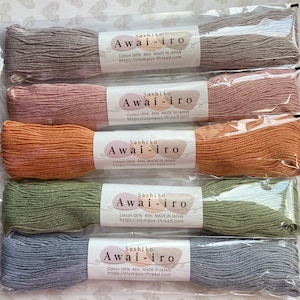 May include: Five spools of Sashiko Awai-iro cotton thread in various colors, including gray, pink, orange, green, and blue. Each spool is labeled with the brand name, thread type, length, and country of origin. The spools are packaged in clear plastic bags.