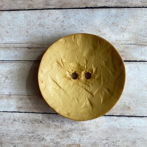 May include: A large, round, gold-colored button with a textured surface and two holes in the center.