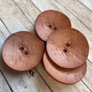 May include: Four large, round, copper-colored buttons with a textured surface. Each button has two holes in the center.