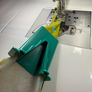 May include: A teal plastic bias tape maker tool folding yellow fabric with a light blue heart pattern, positioned in front of a sewing machine needle and presser foot, with white batting material being fed through the device for assembly.