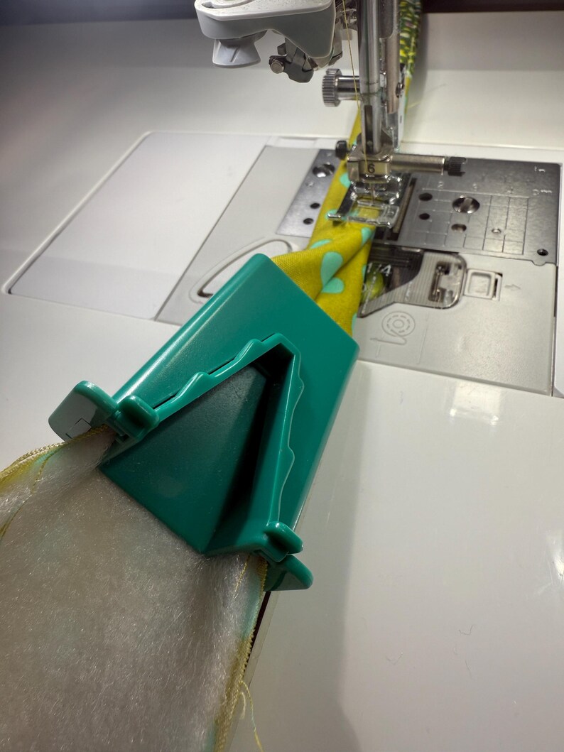 May include: A teal plastic bias tape maker tool folding yellow fabric with a light blue heart pattern, positioned in front of a sewing machine needle and presser foot, with white batting material being fed through the device for assembly.