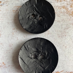 May include: Two black buttons with a textured surface. The buttons have two holes each.