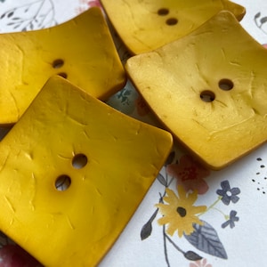 May include: Four gold-colored square buttons with two holes each. The buttons have a textured surface and are arranged in a pile.