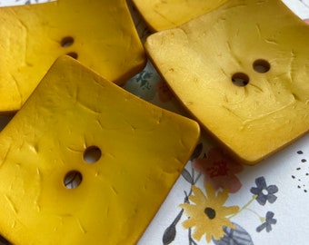May include: Four gold-colored square buttons with two holes each. The buttons have a textured surface and are arranged in a pile.