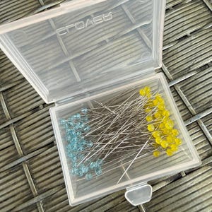 May include: A clear plastic box containing sewing pins. The pins have silver shafts and are topped with blue and yellow beads. The box is open, revealing the pins inside. The word "CPOAEK" is printed on the lid.