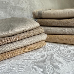 K&ouml;nnte beinhalten: Ein Stapel gefalteter rechteckiger Stoffst&uuml;cke in verschiedenen Beige-, Hellbraun- und Goldbraunt&ouml;nen. Die Textilien weisen eine sichtbare Webstruktur und ausgefranste Kanten an den Seiten auf und sind in zwei Stapeln auf einer wei&szlig;en Fl&auml;che.