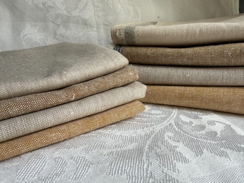 K&ouml;nnte beinhalten: Ein Stapel gefalteter rechteckiger Stoffst&uuml;cke in verschiedenen Beige-, Hellbraun- und Goldbraunt&ouml;nen. Die Textilien weisen eine sichtbare Webstruktur und ausgefranste Kanten an den Seiten auf und sind in zwei Stapeln auf einer wei&szlig;en Fl&auml;che.