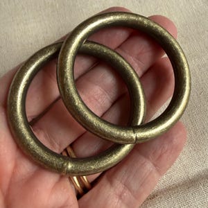 May include: Two circular metal rings with a brushed antique brass finish. The rings feature a smooth, rounded profile and a visible seam where the metal ends meet. The surface has a slightly weathered, textured appearance with subtle color variations.