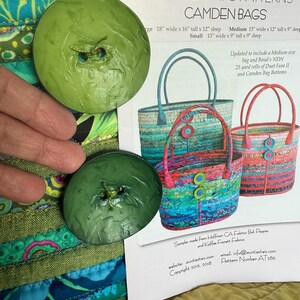 May include: Two large, round, textured green buttons positioned next to a printed sewing pattern titled Aunties Two Patterns Camden Bags. The pattern displays three colorful fabric tote bags with dimensions listed in inches for large, medium, and small sizes.