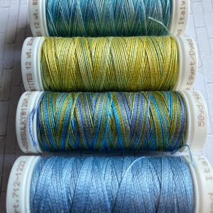 May include: Four spools of embroidery thread in shades of blue, green, and yellow. The spools are labeled with the brand name "Sulky" and the weight of the thread. The thread is made in Italy and Egypt.
