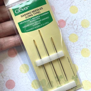 May include: A package of three long Sashiko needles for embroidery. The needles are gold-tipped and packaged in a clear plastic sleeve with a green and yellow label that reads "Clover Sashiko Needles (Long Type)".