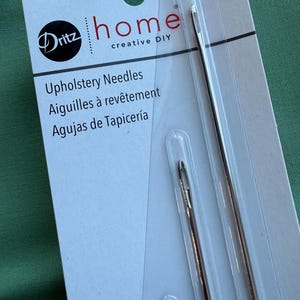 May include: Package of Dritz home upholstery needles. The package is white with the Dritz logo and the words "home creative DIY". The needles are silver and come in a clear plastic sleeve. The package also has text in French and Spanish.