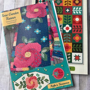 May include: Two quilting pattern books. One titled "Rose Garden Runner" features a large pink rose design on a navy blue background. The other, "Mod Blossoms," showcases a colorful patchwork floral design. Both books are on a light wood grain surface.