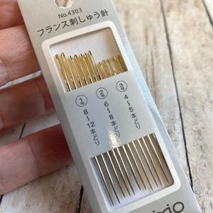 Pode incluir: Um pacote de agulhas de bordar da marca Cosmo. A embalagem cinza claro tem o texto "No.4303" e texto japon&ecirc;s. Cont&eacute;m agulhas douradas nos tamanhos 1, 2 e 3, para bordar.