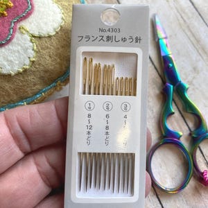 Pode incluir: Um pacote de agulhas de bordar Cosmo com o texto "No.4303" e texto japon&ecirc;s. O pacote cont&eacute;m agulhas douradas de v&aacute;rios tamanhos. Uma tesoura colorida em arco-&iacute;ris &eacute; vis&iacute;vel no fundo.