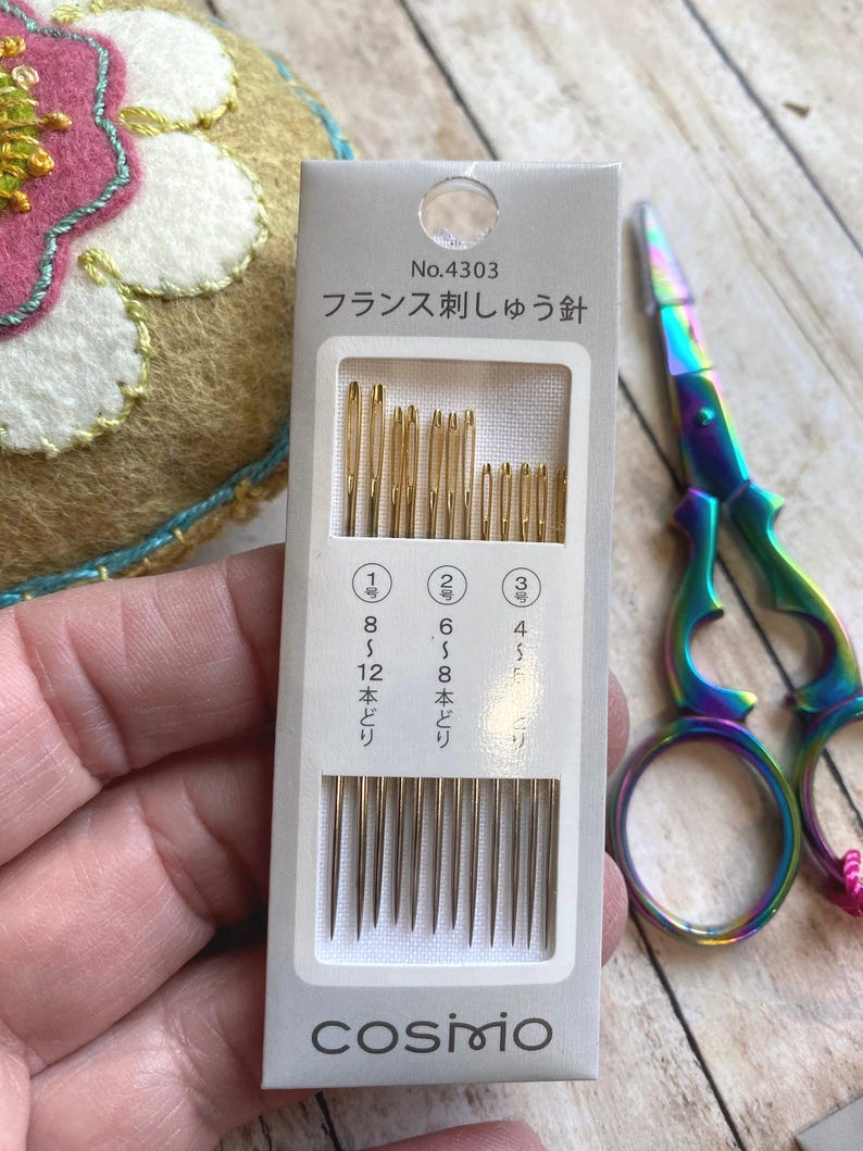 Pode incluir: Um pacote de agulhas de bordar Cosmo com o texto "No.4303" e texto japon&ecirc;s. O pacote cont&eacute;m agulhas douradas de v&aacute;rios tamanhos. Uma tesoura colorida em arco-&iacute;ris &eacute; vis&iacute;vel no fundo.
