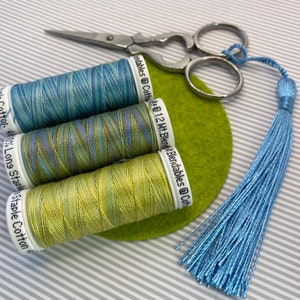 May include: Three spools of embroidery floss in shades of blue, green, and yellow. The floss is arranged on a green felt circle. A pair of silver scissors and a blue tassel are also in the image.