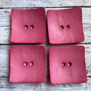 May include: Four square-shaped buttons with a textured surface. The buttons are a deep pink color and have two holes each.
