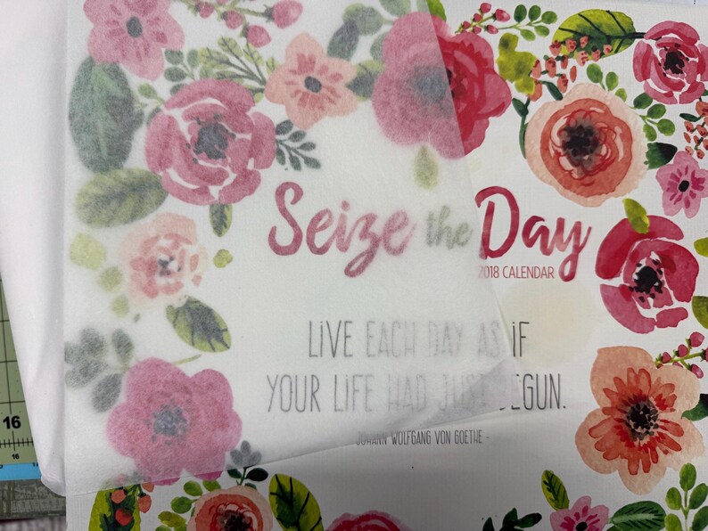 Op de afbeelding: Een kalender voor 2018 met een aquarel bloemenrand in roze, rode en groene tinten. De tekst luidt Seize the Day, Live each day as if your life had just begun en Johann Wolfgang von Goethe. Een doorschijnend vel ligt over de pagina heen.