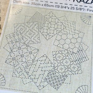 May include: A beige fabric with a starburst design composed of various geometric patterns stitched in black thread. The cloth size is 13 3/4" x 25 5/8".