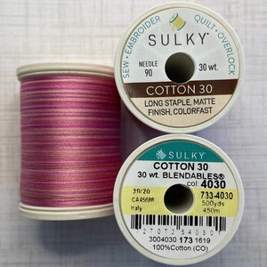 May include: Two spools of Sulky Cotton 30 thread. The spool on the left is pink with thin gold and white stripes. The spool on the right is white with green and yellow text. The text on the spool reads "Sulky Cotton 30 30 wt. Blendables® col: 4030 733-4030 500yds 450m 3004030 173 1619 100%Cotton (CO)"
