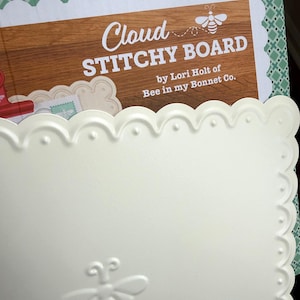 May include: A cream-colored square stitchy board with a scalloped edge and a debossed bee icon in the corner. Behind it sits a product card labeled Cloud STITCHY BOARD by Lori Holt of Bee in my Bonnet Co. with a wood grain background.
