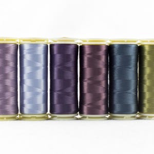May include: Six spools of thread in shades of purple, blue, and green are arranged in a row. The thread is tightly wound on cylindrical spools with yellow bases and tops. The colors range from light lavender to deep teal.