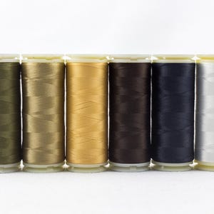 May include: A row of six spools of thread in various colors: olive green, tan, gold, brown, navy blue, and white. The spools are cylindrical with white tops and bottoms, and the thread is tightly wound.