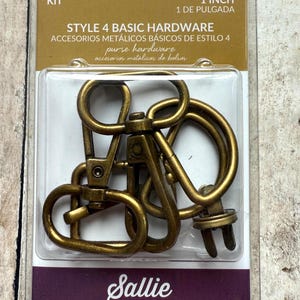 May include: A Sallie Tomato Style 4 Basic Hardware kit containing antique brass-coloured metal swivel lobster clasps and D-rings for purses. The packaging is labelled 1 inch and includes multiple pieces of hardware sealed in a clear plastic blister pack.