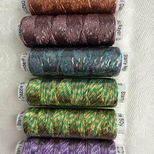 May include: Six spools of WonderFil Dazzle metallic rayon thread stacked vertically. The colors from top to bottom are copper, dark mauve, iridescent blue-green, bright green with gold, olive green with gold, and purple. Each spool has a white label.