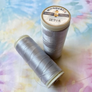Op de afbeelding: Twee spoelen zilvergrijze draad. De spoel bovenop heeft een label met de tekst "Wonderfil InvisaFil  Thread 2 ply  Embroidered Thread  IF718"