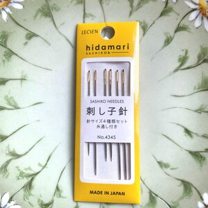 May include: A yellow and white package of sashiko needles, with the brand name "hidamari" and Japanese text. The package contains several needles of varying lengths. The background is a green plate with white daisy designs.