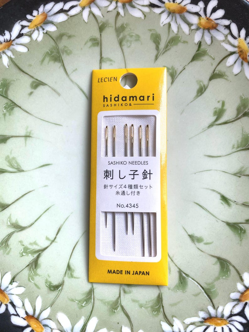 May include: A yellow and white package of sashiko needles, with the brand name "hidamari" and Japanese text. The package contains several needles of varying lengths. The background is a green plate with white daisy designs.