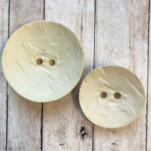 May include: Two cream-colored, oval-shaped buttons with two holes each. The buttons have a textured surface.