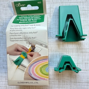 May include: A Clover Fabric Tube Maker kit with two green plastic tools and a box. The box features text in multiple languages and an image of the tool in use. The kit is designed for making Jelly Roll rugs and more.